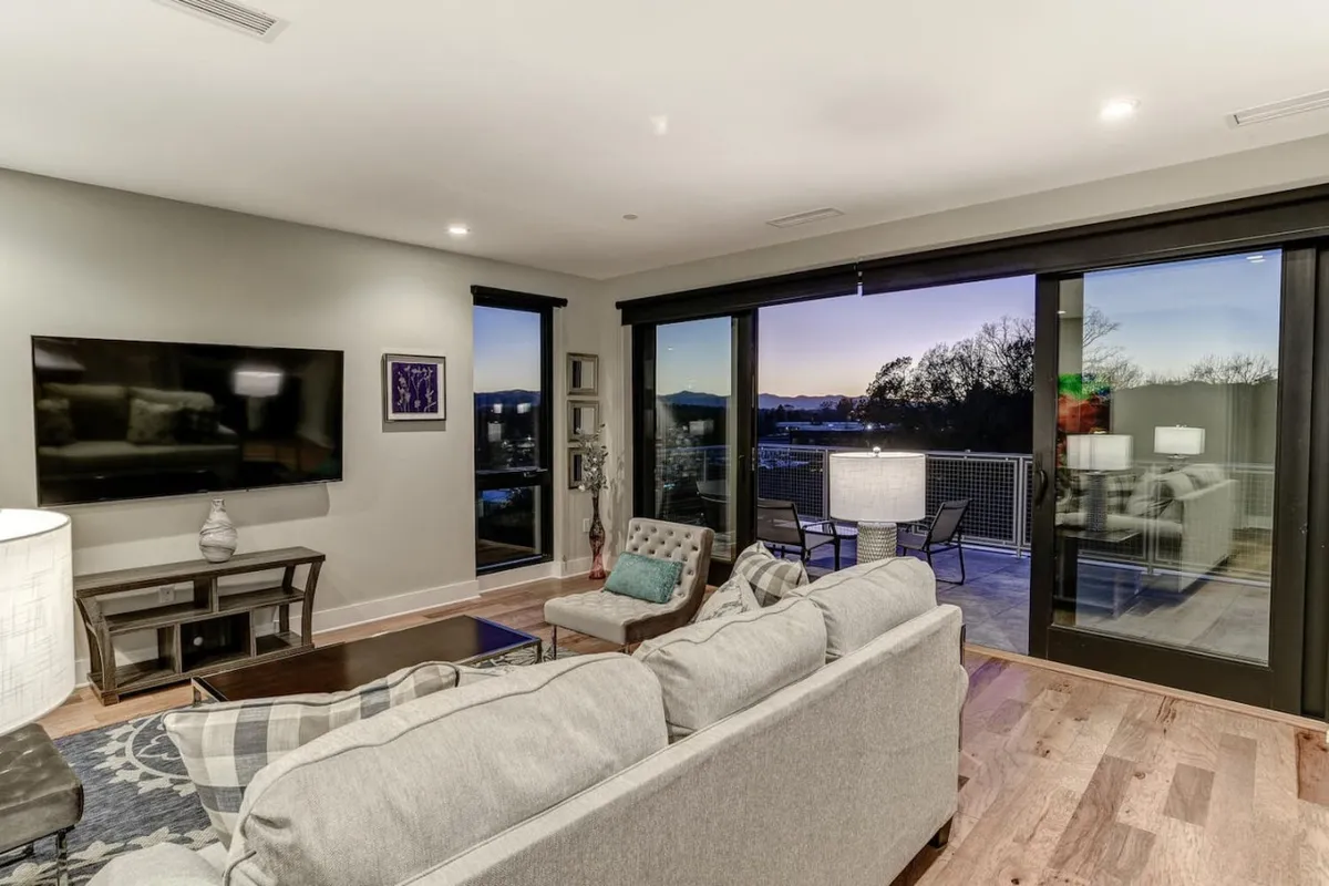 A modern living area with a sofa, large windows offering a striking view, and a dining table set on the balcony. The television and overall décor signal a space equally suited for entertaining guests or cozy movie nights.