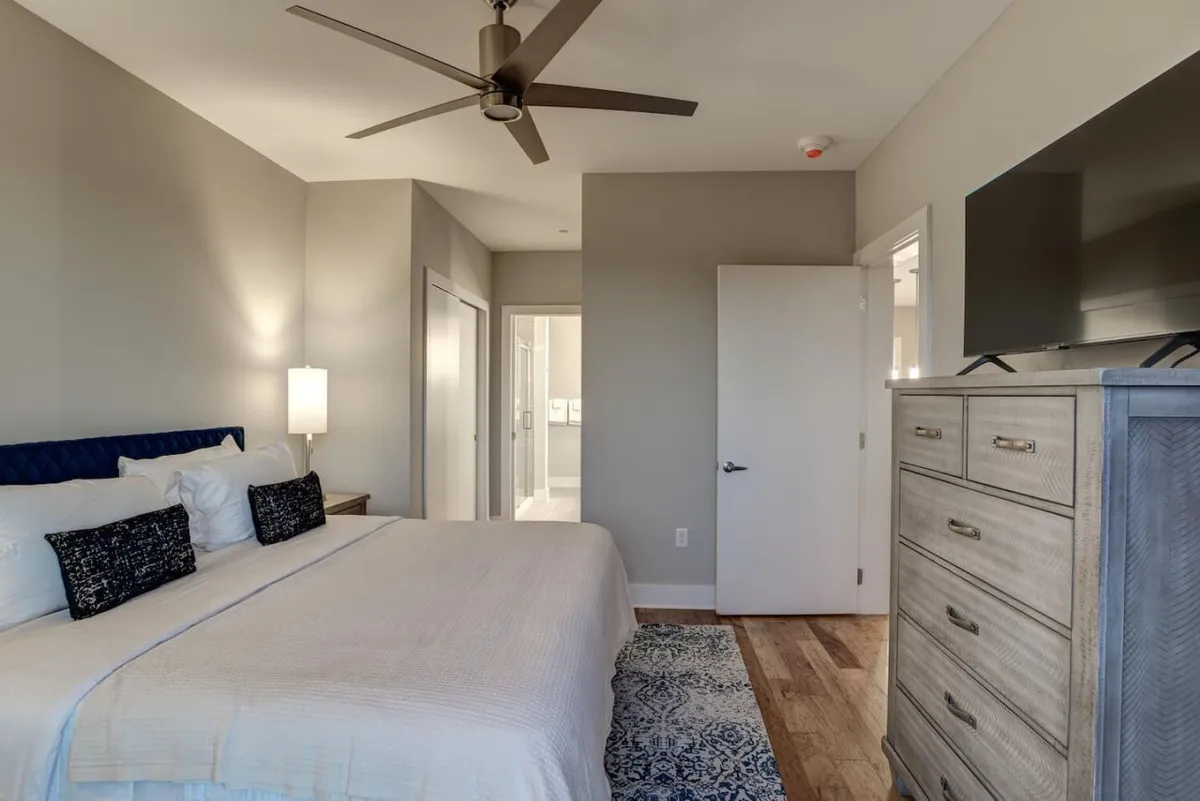 A serene master bedroom featuring a king-size bed adorned with decorative pillows, flanked by a nightstand and a dresser. Natural light streams in, highlighting the cozy reading nook and calm ambiance, ideal for restful nights and peaceful mornings.