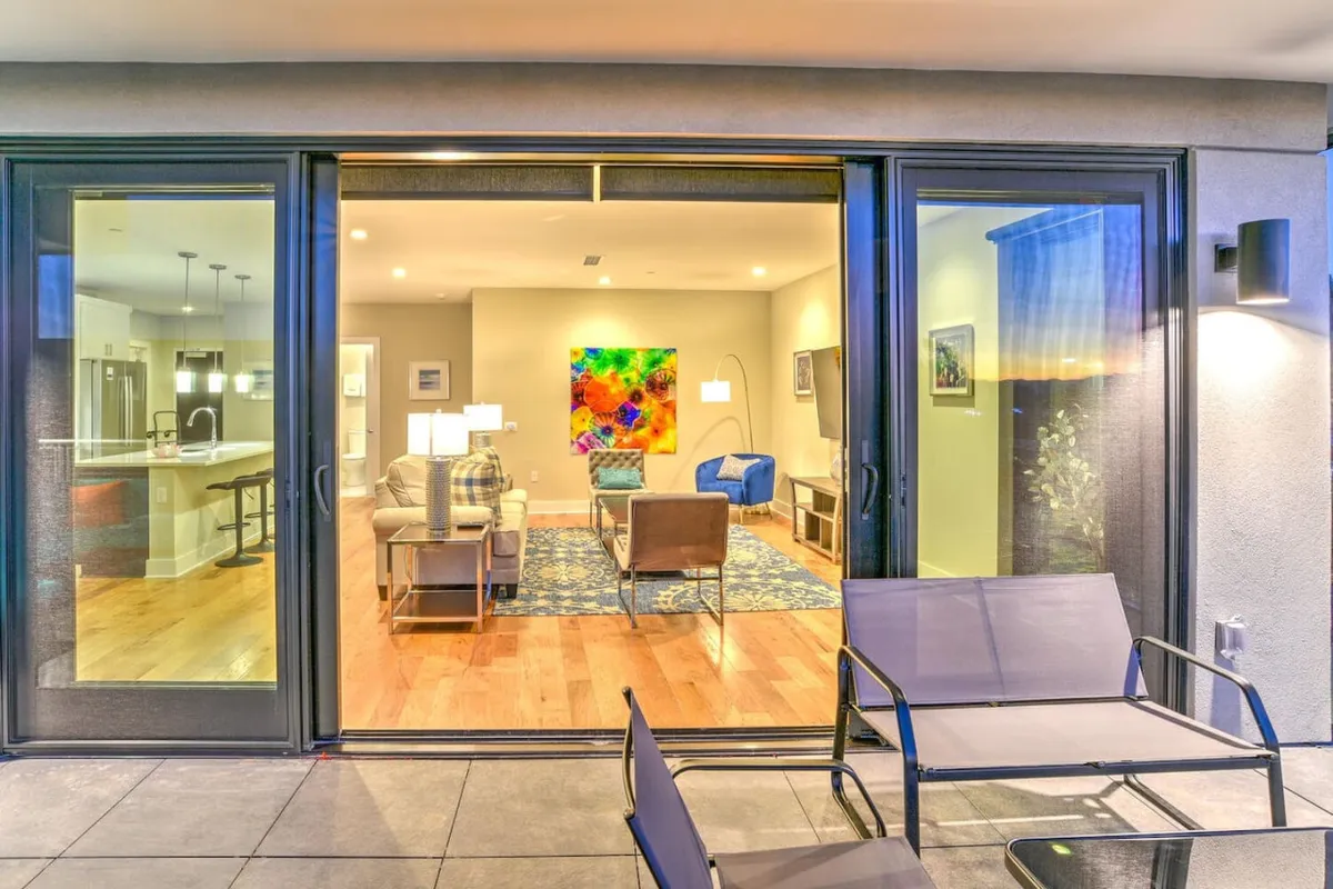 This image captures a vibrant living area featuring colorful artwork on the walls, comfortable sofa seating, and a dining area nearby. The space is illuminated by ample natural light, hinting at large windows and offering glimpses of sunset views from the adjacent patio.