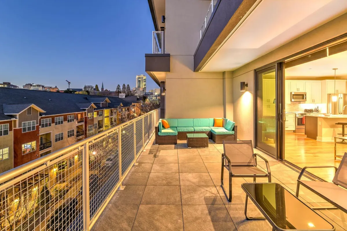 This image captures an inviting balcony seating area overlooking the Asheville city skyline. The photo is taken at dusk, with soft interior lighting contrasting the vibrant cityscape, suggesting a lively downtown atmosphere just outside. The setting invites socializing and taking in city lights, perfectly blending the energy of the city with a private outdoor retreat.