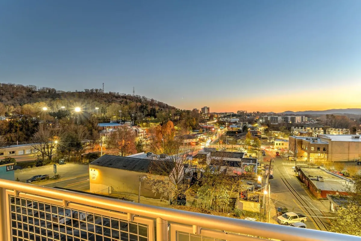 This image captures the Asheville city skyline at twilight, framed by distant hills and illuminated street lights. The building facades and the inviting streets below evoke the lively atmosphere of downtown, hinting at the nearby restaurants, shops, and cultural venues.