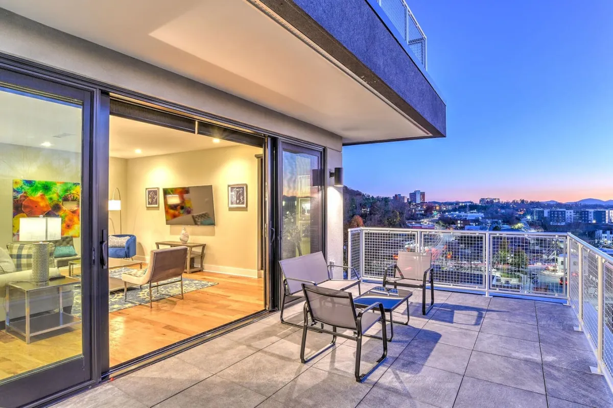 The image showcases an inviting outdoor seating area on a private terrace, with the interior living room visible through large glass doors. The city skyline and rolling Blue Ridge Mountains can be seen in the distance, bathed in the warm glow of sunset. The scene suggests a tranquil yet vibrant atmosphere, perfect for relaxing or entertaining.
