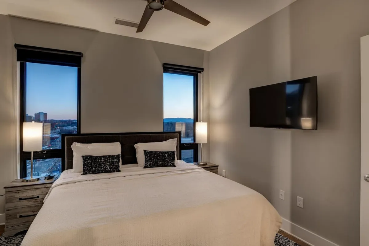 A serene bedroom with large windows framing a calming view, warm lamps casting a gentle light, and a television opposite the bed. The windows hint at an elevated perspective, suggesting the potential for city or mountain vistas from the comfort of the room.