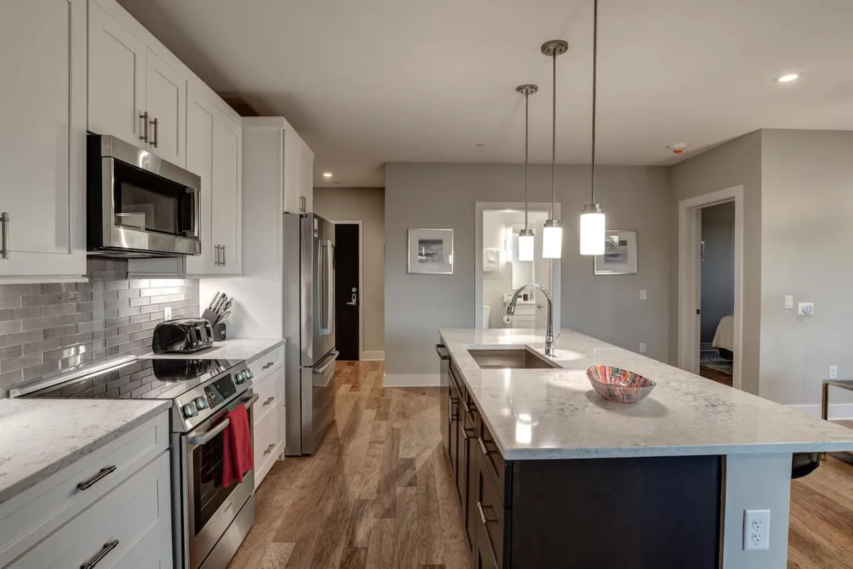 A broad view of the kitchen and dining area, featuring a striking kitchen island, marble (quartz-like) countertop, stainless appliances, and pendant lighting. The scene suggests easy flow between cooking, dining, and socializing.