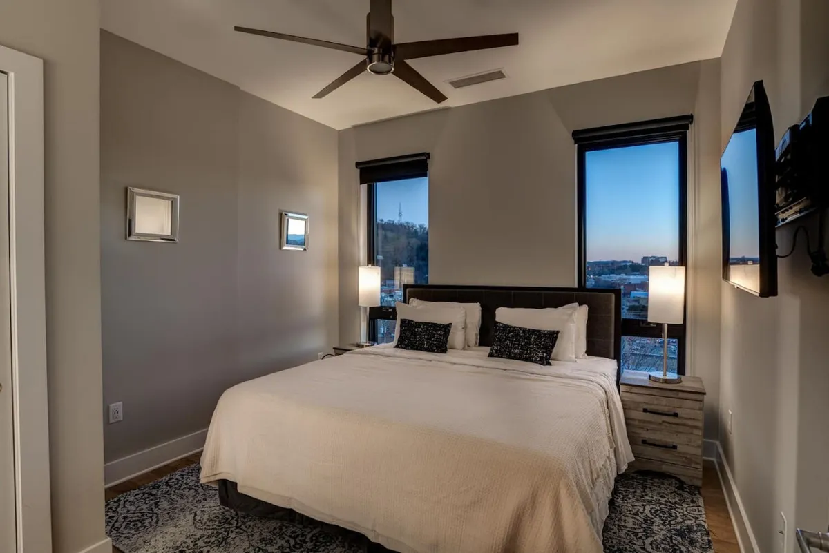 A tranquil bedroom scene featuring a bed positioned near large windows, decorative mirrors reflecting natural light, and a television. The windows offer possible sunset views, reinforcing the idea of relaxation and scenic enjoyment.